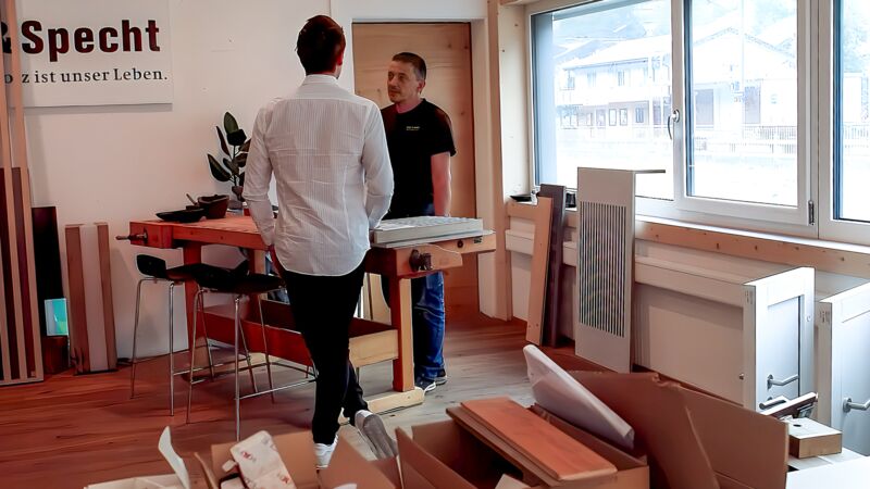 Zwei Männer stehen in einer Werkstatt mit großen Fenstern, einer trägt ein weißes Hemd, der andere ein schwarzes T-Shirt, sie unterhalten sich neben einem Holztisch mit Werkstücken