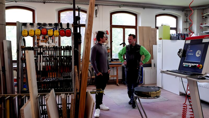 Zwei Männer stehen in einer Werkstatt mit Holzplatten, Werkzeugen und Maschinen, darunter eine CNC-Maschine mit Bildschirm