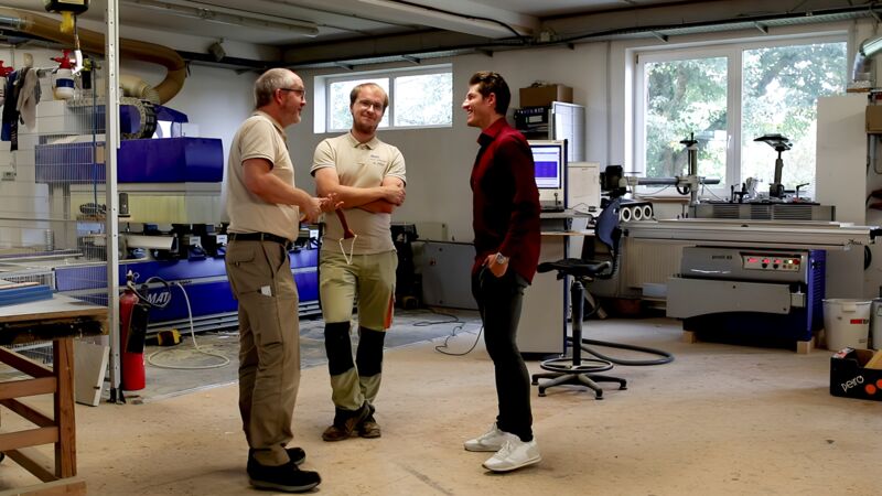 Drei Männer stehen in einer Werkstatt mit Maschinen und besprechen etwas, einer trägt ein rotes Hemd und weiße Schuhe